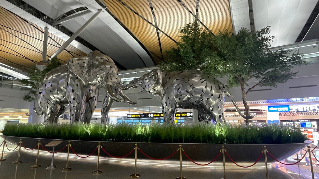 Two large metallic elephant sculptures on display at Bangkok Suvarnabhumi Airport, surrounded by greenery and located near the departure gates under a modern, curved ceiling structure.